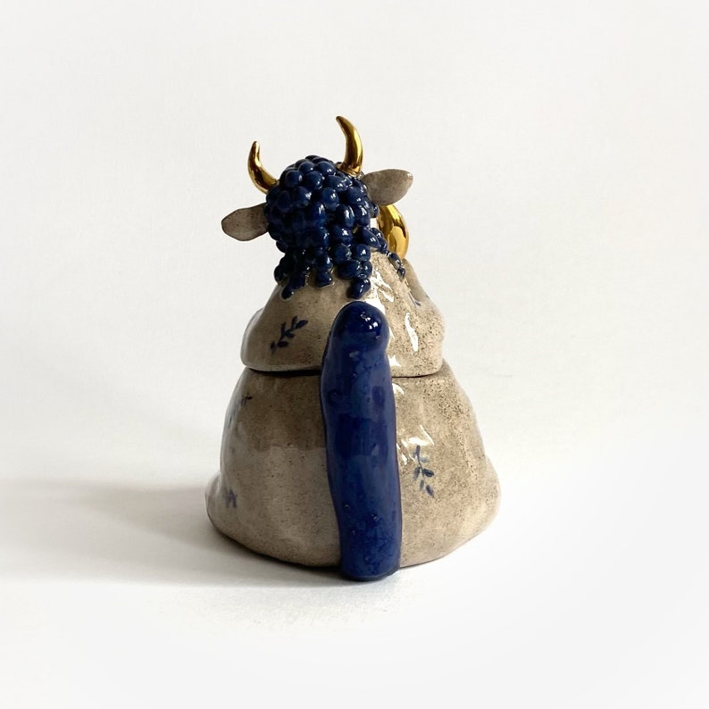 May include: A ceramic cow-shaped container with a lid. The cow has a blue, textured head and tail, with gold-colored horns and ear accents. The body is beige with blue floral accents.