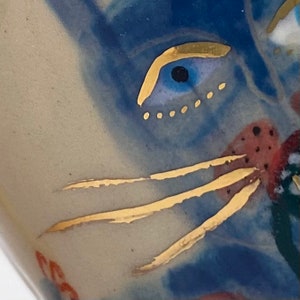 Puede incluir: Primer plano de un ojo de gato pintado en azul y dorado con una ceja y bigotes dorados. El ojo est&aacute; rodeado de un fondo azul y blanco.