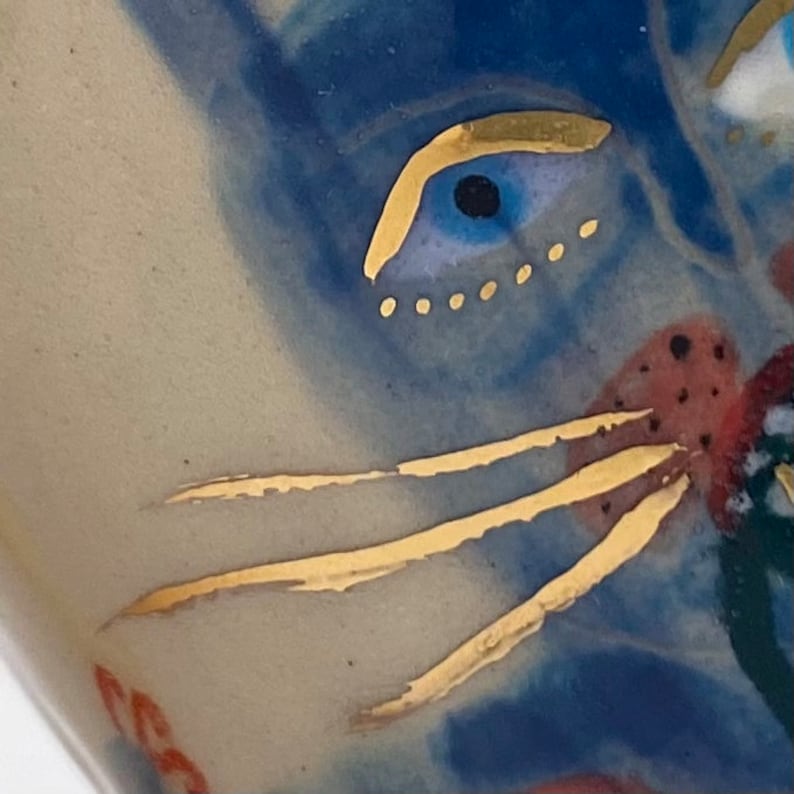 Puede incluir: Primer plano de un ojo de gato pintado en azul y dorado con una ceja y bigotes dorados. El ojo est&aacute; rodeado de un fondo azul y blanco.