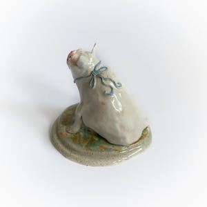 May include: A ceramic pig figurine, off-white with pink spots, sits on a round base with green and tan accents. The pig has a blue ribbon around its neck and is looking upwards. A unique decorative item.