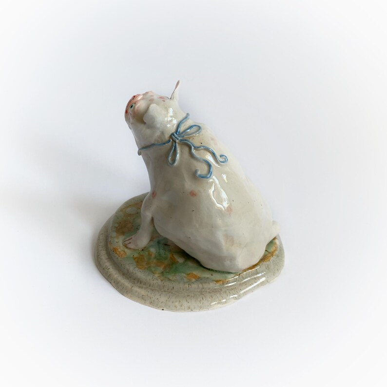 May include: A ceramic pig figurine, off-white with pink spots, sits on a round base with green and tan accents. The pig has a blue ribbon around its neck and is looking upwards. A unique decorative item.