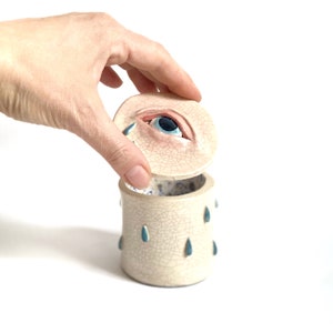 Könnte beinhalten: Eine weiße Keramikdose mit einem Deckel in Form eines weinenden Auges. Der Deckel hat ein blaues Auge mit einer schwarzen Pupille und Wimpern. Die Dose ist mit blauen Tränentropfen verziert.