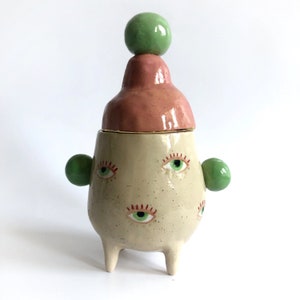 Puede incluir: Un tarro de cerámica con tapa y tres patas. El tarro tiene una base blanca con ojos verdes y rojos pintados. La tapa es rosa con una bola verde en la parte superior.