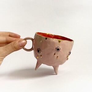 May include: Pink ceramic mug with three legs and gold accents. The mug has three eyes with blue and black details and gold teardrop shapes.