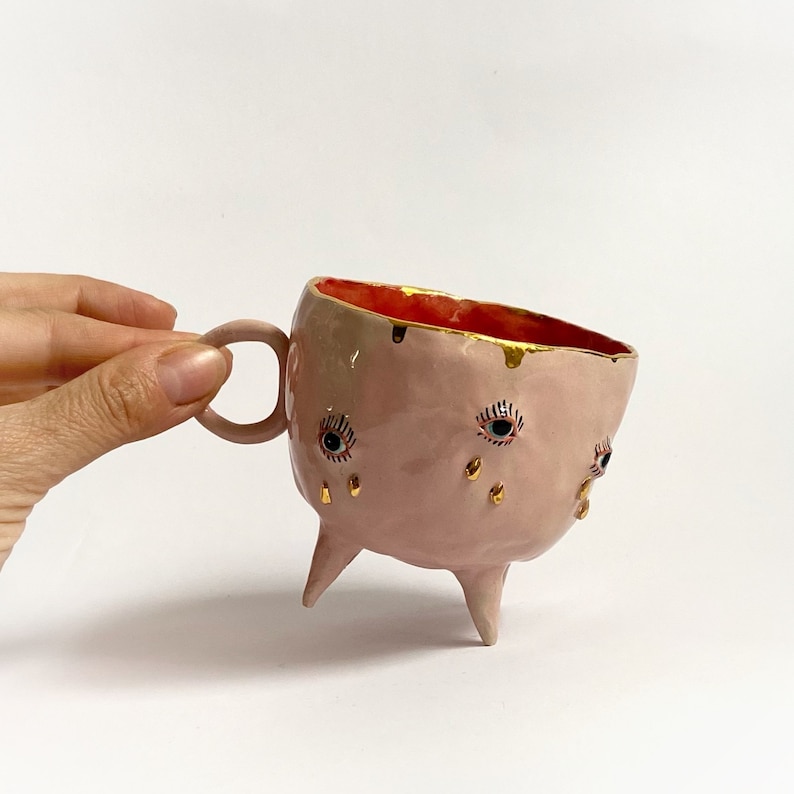 May include: Pink ceramic mug with three legs and gold accents. The mug has three eyes with blue and black details and gold teardrop shapes.