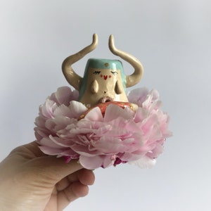 Puede incluir: Una figurita de cerámica de una persona con los brazos extendidos, que lleva un sombrero azul claro y está sentada en un lecho de pétalos de flores rosas. La figurita tiene una cara pintada con los ojos cerrados y un corazón rojo en el pecho.