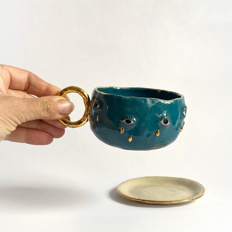 K&ouml;nnte beinhalten: Eine teeblaue Keramiktasse mit einem goldenen Ringgriff und goldenen Augen mit goldenen Tr&auml;nentropfen. Die Teetasse steht auf einer wei&szlig;en Untertasse mit goldenem Rand.