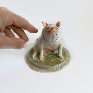 May include: A ceramic bulldog figurine in light pink and white. The dog has a blue collar and sits on a round, green and grey base. The figurine is detailed with painted features, showcasing a unique artistic style.