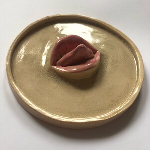 Pode incluir: Um prato de cer&acirc;mica com uma escultura de l&aacute;bios rosados em relevo e esmaltada no centro. O prato &eacute; bege com um esmalte craquelado.