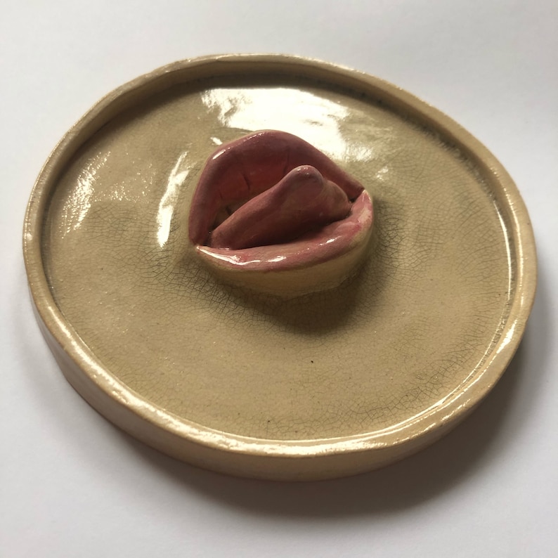 Pode incluir: Um prato de cer&acirc;mica com uma escultura de l&aacute;bios rosados em relevo e esmaltada no centro. O prato &eacute; bege com um esmalte craquelado.