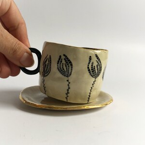 May include: A ceramic teacup and saucer set with a black floral design and gold trim. The cup has a unique, slightly asymmetrical shape.