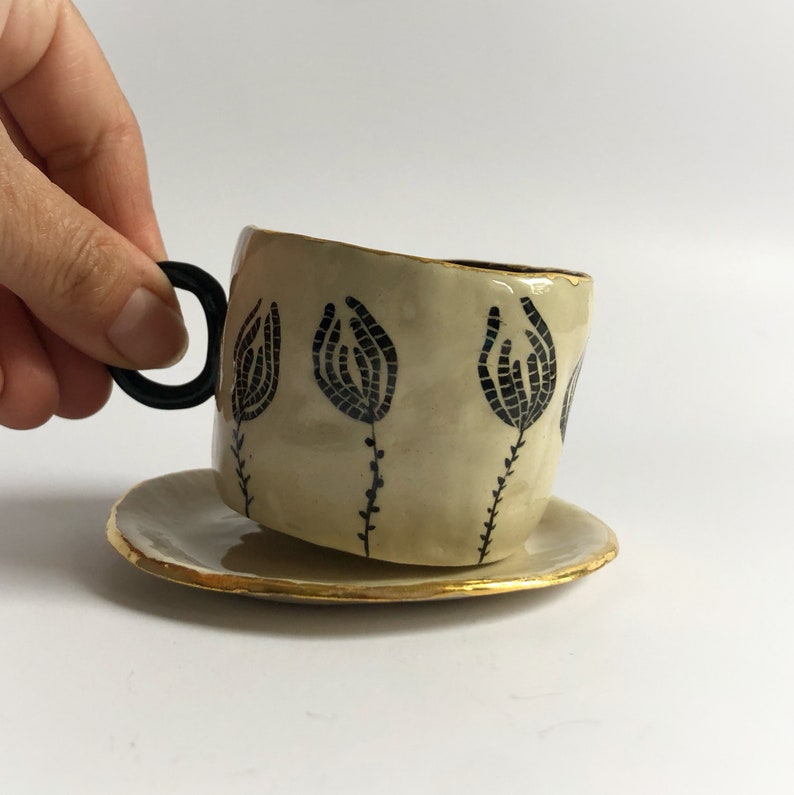 May include: A ceramic teacup and saucer set with a black floral design and gold trim. The cup has a unique, slightly asymmetrical shape.