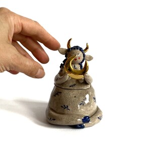 May include: A ceramic figurine of a cow-like creature with closed eyes, holding a gold crescent moon. The figurine is beige with blue floral accents and gold horns. The cow is wearing a blue braided hair. The figurine is on a white background.