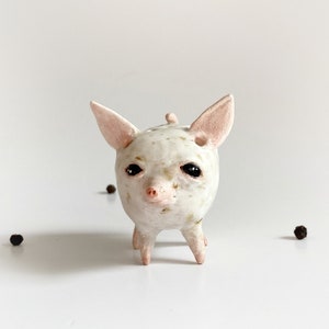 Puede incluir: Una figurita de cer&aacute;mica blanca de un cerdo con ojos negros y una nariz rosa. El cerdo tiene un esmalte blanco y marr&oacute;n moteado.