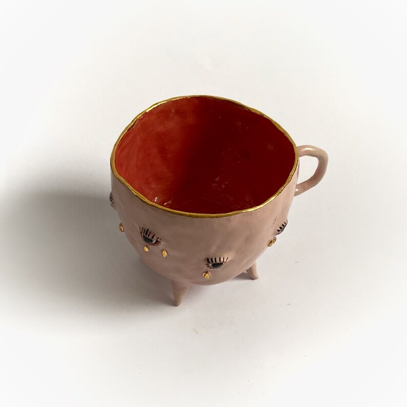 May include: A pink ceramic cup with a red interior and gold rim. The cup has three legs and is decorated with black and gold eyes.