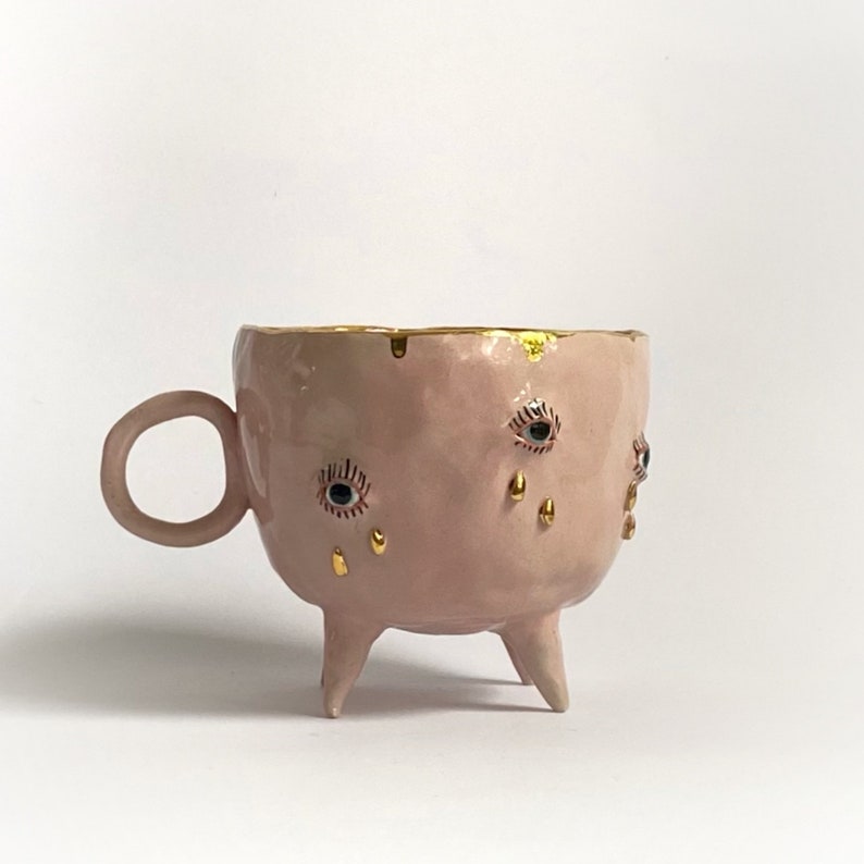 May include: A pink ceramic mug with three legs and gold accents. The mug has three eyes with gold eyelashes and gold teardrop details.