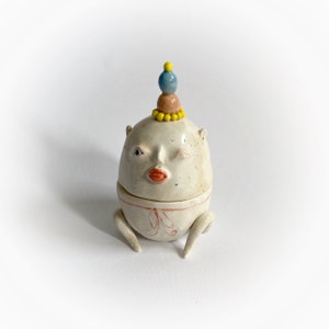 Może przedstawiać: Biała ceramiczna figurka z mrugającym okiem, czerwonymi ustami i żółto-niebieską koroną. Figurka ma dwie małe nogi i stoi na białym tle.