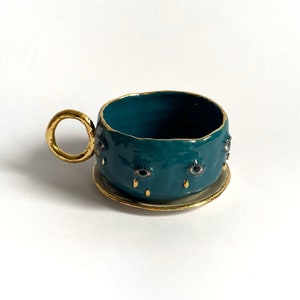 K&ouml;nnte beinhalten: Eine teeblau gef&auml;rbte Keramiktasse mit goldenem Henkel und Untertasse. Die Tasse ist mit mehreren goldger&auml;nderten Augen mit goldenen Tr&auml;nenakzenten verziert.