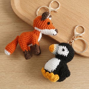 May include: Two crochet animal keychains: an orange fox with white and brown accents and a black, white, and yellow penguin. Both keychains have gold-colored metal clasps.