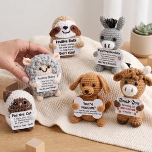 Op de afbeelding: Een verzameling van zes kleine gehaakte dierenfiguren, waaronder een luiaard, ezel, schaap, kat, hond en koe. Elk figuur houdt een wit kaartje vast met gedrukte tekst met positieve affirmaties en woordspelingen zoals Positive Sloth, Positive Donkey, Positive Sheep, Positive Cat, You're PAWSOME en Thank You for being the MOO-TIVATION in my life.