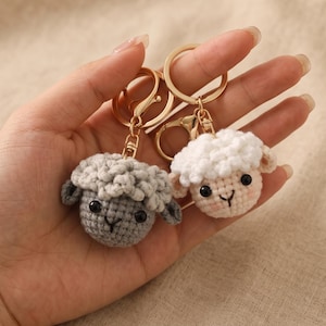 May include: Two crochet sheep keychains. One is gray, the other is white with pink cheeks. Both have black eyes and gold key rings. The sheep have a fluffy wool texture and are handmade.