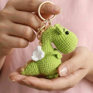 May include: A green crocheted dragon keychain with white wings and a gold-colored keyring. The dragon has a light brown spine and black eyes. The keychain is held in open hands.