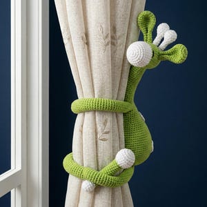 May include: A whimsical, green crocheted giraffe curtain tieback wraps around a beige curtain. The giraffe has white accents on its head and body. The curtain has a subtle leaf pattern. The background is a dark blue.