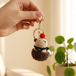 May include: A handmade crochet keychain featuring a small bear-like figure in a brown basket. The bear has a cream-colored body, a black scarf, and a red cherry on its head. The keychain has a gold-colored clasp.