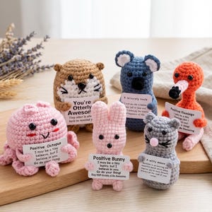 May include: A collection of six small crocheted animals including a pink octopus, brown otter, blue koala, orange flamingo, pink bunny, and gray mouse. Each figure holds a small white card with printed text and a positive message.
