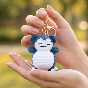 Peut inclure: Porte-clés crocheté représentant un personnage bleu et blanc, avec un porte-clés et un fermoir dorés. La peluche faite à la main a un corps rond, de petits bras et jambes, et une expression endormie. Le porte-clés est tenu dans les mains d'une personne sur un fond vert flou.