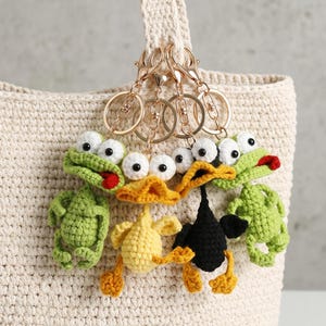 May include: Crocheted keychains featuring a frog, a duck, and a black bird. The keychains are attached to gold-colored rings and clasps. The frog is green with a red tongue, the duck is yellow, and the bird is black. The keychains are hanging on a cream-colored bag.