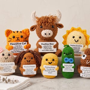 May include: A collection of eight small, crocheted plush figures including a cat, a highland cow, a sun, two potatoes, a pickle, and a brown character, each holding a white card with printed motivational text and a specific character name.