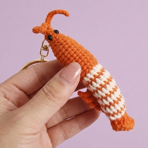 May include: A handmade orange and white crochet shrimp keychain with black eyes and a gold-colored clasp. The shrimp has a segmented body with a white and orange striped pattern. The keychain is a fun, whimsical accessory.