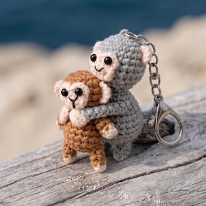 Può includere: Un portachiavi all'uncinetto con una figura di scimmia grigia più grande che abbraccia una figura di scimmia marrone più piccola. Entrambe le scimmie hanno il viso beige chiaro, occhi a perla neri e bocche ricamate. Una catena in metallo argentato con moschettone è attaccata alla scimmia grigia.