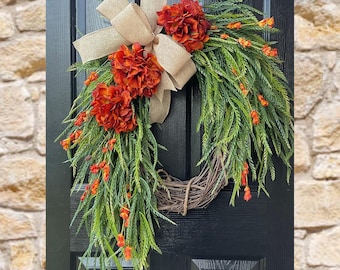 May include: A decorative wreath featuring a woven grapevine base, adorned with vibrant orange-red flowers, green foliage, and small orange accents. A large burlap bow adds a rustic touch. The wreath hangs on a black door.