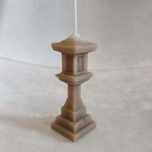 Puede incluir: Una vela de color beige con forma de pagoda japonesa. La vela tiene una base cuadrada, un pedestal escalonado y un techo de varios niveles. Una mecha blanca sobresale de la parte superior. El artículo es decorativo.