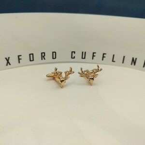 May include: Two gold-coloured stag head cufflinks. The cufflinks are displayed on a white surface, with the words "OXFORD CUFFLINKS" printed in black.