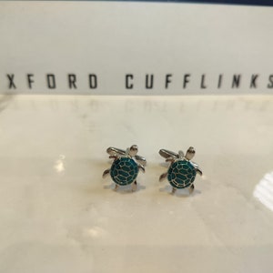 May include: A pair of silver cufflinks featuring a green turtle design. The cufflinks are displayed on a white surface with the text "OXFORD CUFFLINKS" visible in the background.