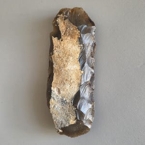 May include: A hand-knapped flint tool with a light brown and gray color. The tool has a rough, textured surface with a chipped edge, and a smooth, polished area. The tool is likely an ancient artifact.