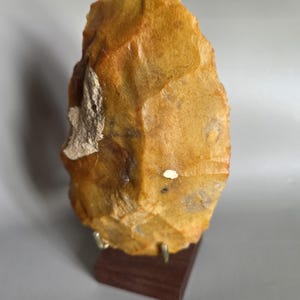 May include: A hand-knapped, golden-brown flint stone tool with a pointed top and textured, chipped edges. The stone features a small, rough, light-colored patch on its left side and is displayed upright on a dark brown wooden base stand.