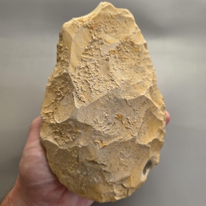 Puede incluir: Una herramienta de piedra tallada a mano con forma de gota, que presenta una superficie texturizada de color tostado con cicatrices de lascas visibles y bordes afilados e irregulares. El objeto tiene un aspecto rugoso y poroso.