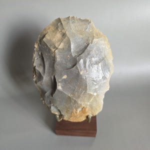 Può includere: Un'ascia a mano in selce grigia di forma ovale con bordi scheggiati visibili e una superficie ruvida, montata in verticale su una piccola base rettangolare in legno marrone scuro con due sottili perni metallici che sostengono il reperto.