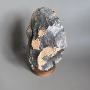 May include: A dark gray, roughly shaped stone tool, possibly a hand axe, with lighter tan-colored patches. The stone is mounted on a small, round wooden base, showcasing its primitive form and texture.