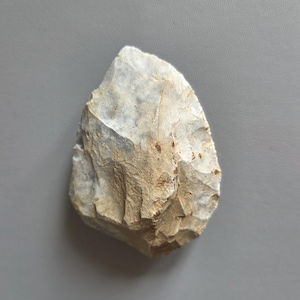 Könnte beinhalten: Ein handgeformtes Steinwerkzeug in Hellgrau und Beige. Die Oberfläche ist rau und die Kanten sind abgebrochen, was auf eine Verwendung zum Schneiden oder Schaben hindeutet. Das Artefakt ist wahrscheinlich aus der Altsteinzeit und ist eine Faustkeil.