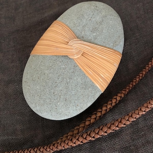May include: An oval grey stone with a woven tan band tied around it. A braided brown leather cord is also visible. The woven material is tied in the centre of the stone.