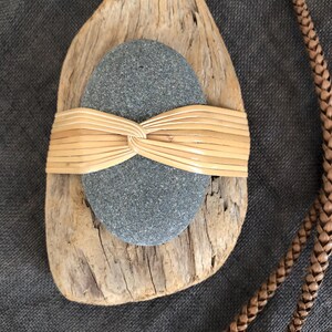 Peut inclure: Pendentif artisanal composé d'une pierre grise enveloppée de matériau tissé beige, posé sur un morceau de bois flotté. Un cordon en cuir tressé est visible sur le côté droit. L'ensemble dégage une esthétique rustique et naturelle.