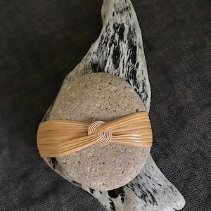 May include: A round, grey stone with a woven, light brown band tied around it in a knot. The stone is sitting on a piece of driftwood.