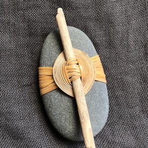 May include: A grey, oval stone with a woven, circular design and a wooden stick across it. The woven design is made of thin, light brown strips. The stick is light brown and angled across the stone.