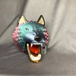 Könnte beinhalten: Eine 3D-gedruckte Wolfskopfskulptur mit blauer, rosa und goldener Lackierung. Der Wolf hat den Mund offen, wodurch eine rote Zunge und weiße Zähne sichtbar werden.