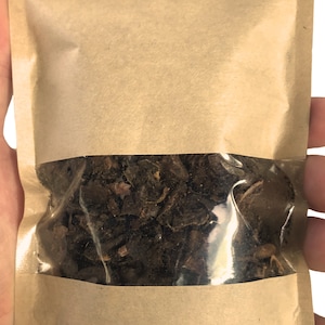 以下が含まれることがあります： 中央に透明なプラスチック窓が付いた長方形の茶色の紙袋。袋の中には、不規則な形の暗褐色の乾燥した有機物が詰められており、透明な窓からその質感のある脆そうな様子が見えています。